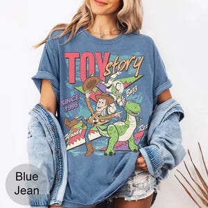 May include: Blue t-shirt featuring a graphic of Toy Story characters Woody, Buzz, and Rex, with the text "TOY STORY" and "SINCE 1995". The shirt has rolled-up sleeves and a vintage style.