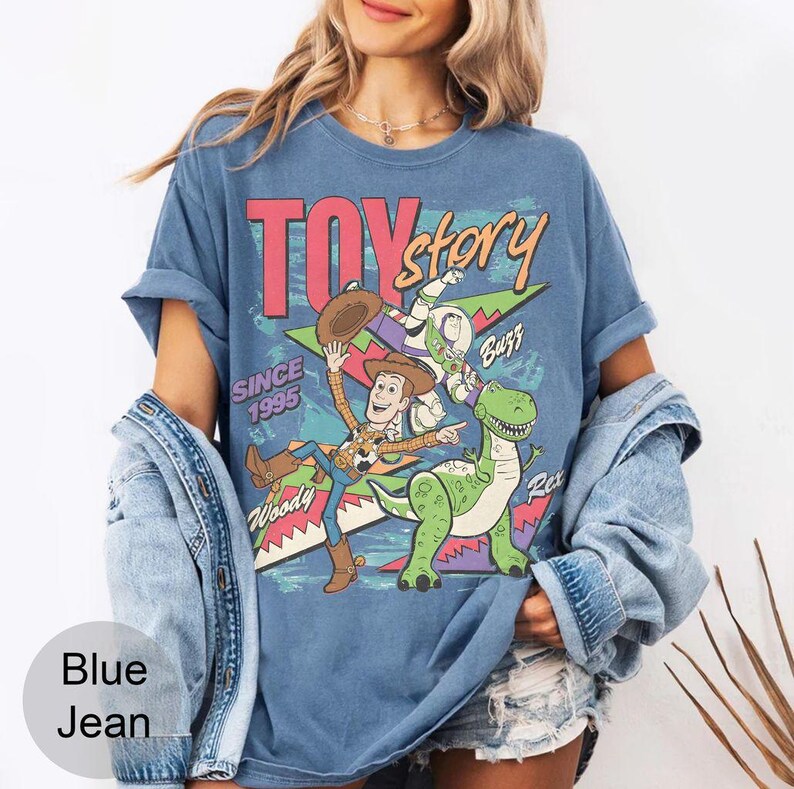 May include: Blue t-shirt featuring a graphic of Toy Story characters Woody, Buzz, and Rex, with the text "TOY STORY" and "SINCE 1995". The shirt has rolled-up sleeves and a vintage style.