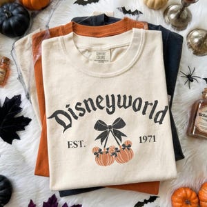 May include: A cream-colored t-shirt featuring the black text Disneyworld, the text EST. 1971, and a graphic of two orange pumpkins with black bows. The shirt has a crew neckline and a Comfort Colors brand label printed on the inside of the collar.