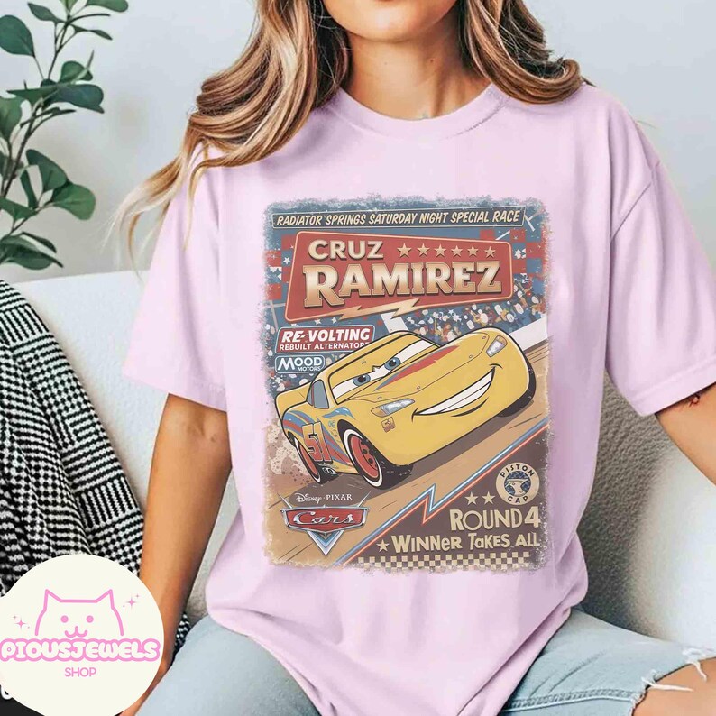 May include: A light pink t-shirt featuring a graphic from the Disney Pixar Cars movie. The graphic includes the text "CRUZ RAMIREZ" and a yellow race car. The shirt also has the text "RADIATOR SPRINGS SATURDAY NIGHT SPECIAL RACE" and "ROUND 4 WINNER TAKES ALL".