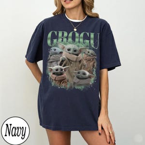 Pode incluir: Camiseta azul marinho com um gr&aacute;fico de Grogu, tamb&eacute;m conhecido como Baby Yoda, e a palavra "GROGU" em verde. O design inclui v&aacute;rias imagens de Grogu. A camiseta &eacute; um estilo confort&aacute;vel e casual.