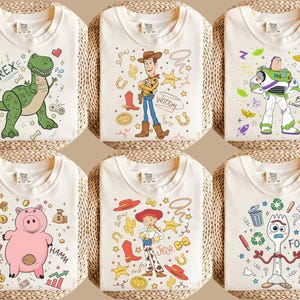 May include: Six off-white t-shirts with colorful cartoon character designs. The designs include a green dinosaur, Woody, Buzz Lightyear, Hamm the pig, Jessie, and Forky. Each shirt is folded and displayed on a woven surface.
