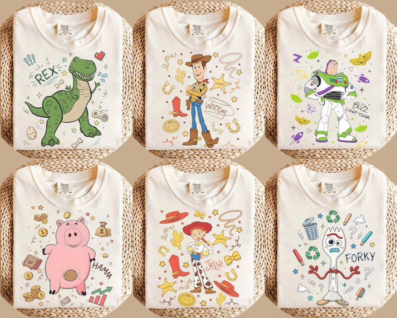 May include: Six off-white t-shirts with colorful cartoon character designs. The designs include a green dinosaur, Woody, Buzz Lightyear, Hamm the pig, Jessie, and Forky. Each shirt is folded and displayed on a woven surface.