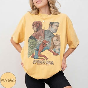 May include: Mustard-coloured t-shirt featuring a Spider-Man graphic with other Marvel characters. The graphic includes the text "SPIDER-MAN BRAND NEW DAY". The shirt is a casual wear item.