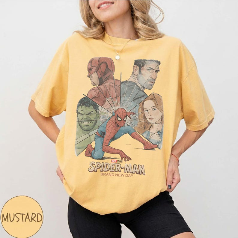 May include: Mustard-coloured t-shirt featuring a Spider-Man graphic with other Marvel characters. The graphic includes the text "SPIDER-MAN BRAND NEW DAY". The shirt is a casual wear item.