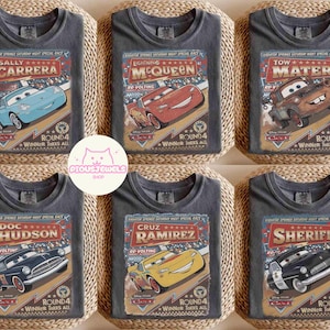 May include: Six gray t-shirts featuring characters from the Disney Cars movie. Each shirt displays a different character: Sally Carrera, Lightning McQueen, Tow Mater, Doc Hudson, Cruz Ramirez, and Sheriff. The shirts have a vintage racing theme with text and graphics.