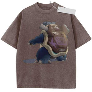 May include: A brown acid-wash short-sleeve t-shirt featuring a graphic of a blue creature with a large brown shell on its back. The shell has two metallic cannons and purple markings. A white rectangular clothing tag is attached to the collar.
