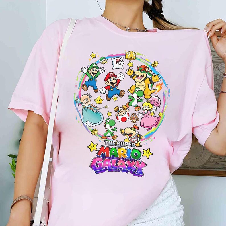 K&ouml;nnte beinhalten: Hellrosa T-Shirt mit einem farbenfrohen Grafikdesign von Charakteren aus Super Mario Galaxy. Das Design zeigt Mario, Luigi, Prinzessin Peach, Yoshi und Bowser, umgeben von Sternen und einem Regenbogen.