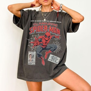 K&ouml;nnte beinhalten: Dunkelgraues T-Shirt mit einem Comic-Design von Spider-Man. Das Design enth&auml;lt den Text "the AMAZING SPIDER-MAN" in Rot und Wei&szlig; sowie weitere Comic-Elemente. Das Shirt hat kurze &Auml;rmel und eine lockere Passform.