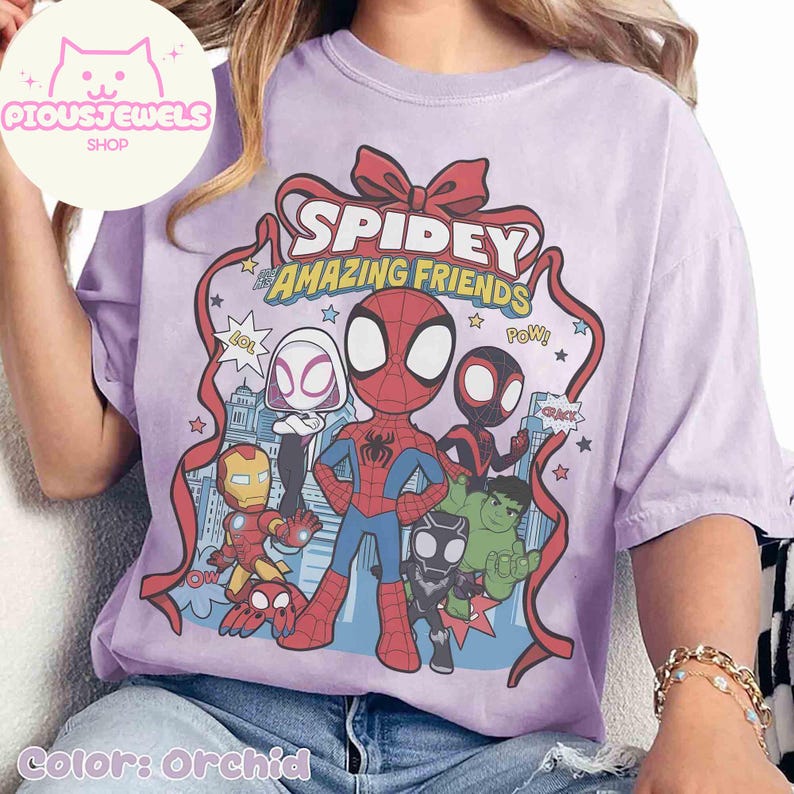 Puede incluir: Camiseta color orqu&iacute;dea con un gr&aacute;fico de Spidey y sus incre&iacute;bles amigos. El dise&ntilde;o incluye a Spider-Man, Spider-Gwen y otros personajes. El texto "Spidey & Amazing Friends" se muestra en un dise&ntilde;o de cinta roja. La camiseta es de corte holgado.