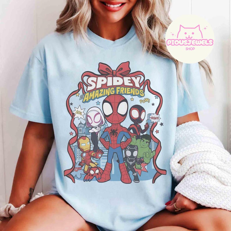 Puede incluir: Camiseta azul claro con un dise&ntilde;o de Spidey y sus Incre&iacute;bles Amigos. El dise&ntilde;o incluye a Spider-Man, Iron Man, Hulk y otros personajes. El texto "Spidey Amazing Friends" se muestra en una cinta roja.