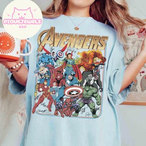 Puede incluir: Una camiseta azul claro con un gran estampado gr&aacute;fico de varios personajes de c&oacute;mics de Marvel, incluidos Spider-Man, Hulk y Capit&aacute;n Am&eacute;rica, con la palabra "AVENGERS" en letras doradas grandes en la parte superior y varias firmas en tinta negra.