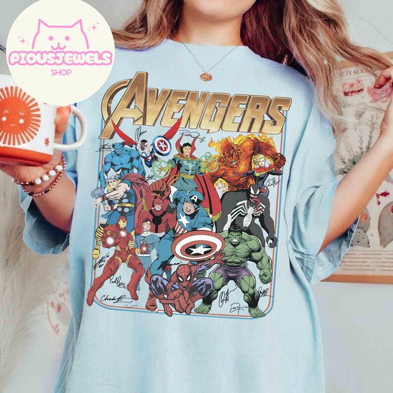 Puede incluir: Una camiseta azul claro con un gran estampado gr&aacute;fico de varios personajes de c&oacute;mics de Marvel, incluidos Spider-Man, Hulk y Capit&aacute;n Am&eacute;rica, con la palabra "AVENGERS" en letras doradas grandes en la parte superior y varias firmas en tinta negra.