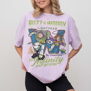 Może przedstawiać: Jasnofioletowa koszulka z grafiką Buzza Astrala i Chudego z Toy Story. Tekst "Buzz & Woody Lightyear" i "To Infinity and Beyond" jest wyświetlany w kolorze zielonym i białym.