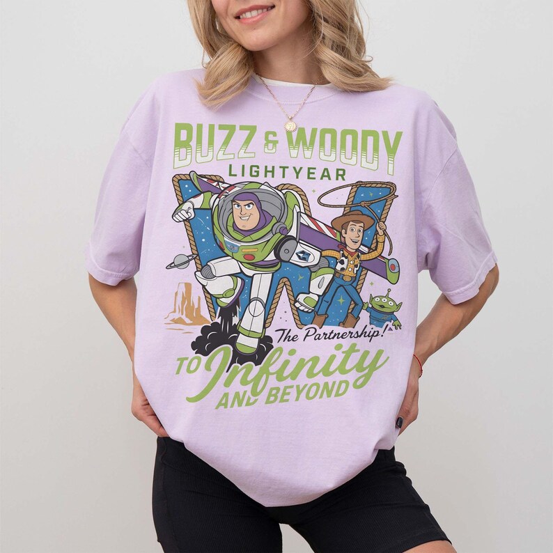 Może przedstawiać: Jasnofioletowa koszulka z grafiką Buzza Astrala i Chudego z Toy Story. Tekst "Buzz & Woody Lightyear" i "To Infinity and Beyond" jest wyświetlany w kolorze zielonym i białym.