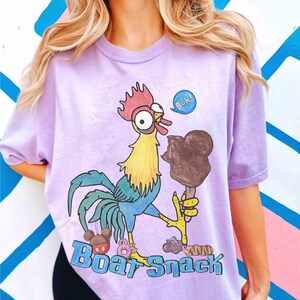 May include: A light purple t-shirt featuring a cartoon rooster holding a chocolate ice cream bar. The rooster has red, yellow, blue, and green coloring. The text "Bok!" is in a blue speech bubble. The words "Boat Snack" are at the bottom.
