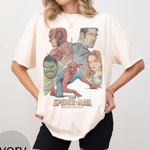 May include: Ivory t-shirt with a graphic of Spider-Man and other Marvel characters. The design includes the text "Spider-Man Brand New Day". The shirt has short sleeves and a crew neck.