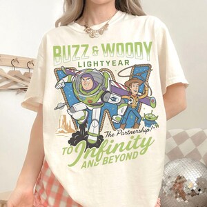 Może przedstawiać: Kremowa koszulka z grafiką Buzza Astrala i Chudego z Toy Story. Projekt zawiera tekst "Buzz & Woody Lightyear" i "To Infinity and Beyond" w kolorze zielonym. Koszulka ma kr&oacute;tkie rękawy.