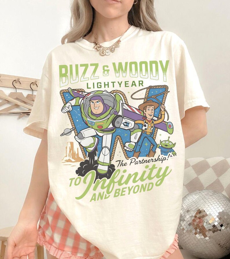 Może przedstawiać: Kremowa koszulka z grafiką Buzza Astrala i Chudego z Toy Story. Projekt zawiera tekst "Buzz & Woody Lightyear" i "To Infinity and Beyond" w kolorze zielonym. Koszulka ma kr&oacute;tkie rękawy.