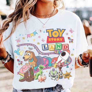 May include: White t-shirt with a vibrant "Toy Story Land" design. The graphic includes cartoon characters, rockets, and a train, with the words "Toy Story" and "Land" in bold letters. The shirt has a crew neck and short sleeves.