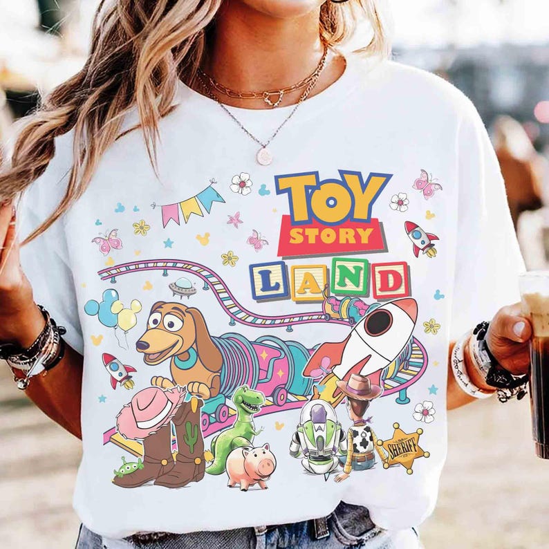 May include: White t-shirt with a vibrant "Toy Story Land" design. The graphic includes cartoon characters, rockets, and a train, with the words "Toy Story" and "Land" in bold letters. The shirt has a crew neck and short sleeves.