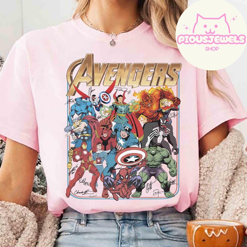 Puede incluir: Una camiseta de manga corta de color rosa claro con un gran estampado gr&aacute;fico de varios personajes de c&oacute;mics de Marvel. La palabra AVENGERS est&aacute; impresa en dorado en la parte superior del dise&ntilde;o, que incluye figuras y firmas.