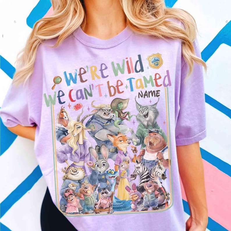 Puede incluir: Una camiseta de color lila claro con un gr&aacute;fico colorido de varios personajes de animales de dibujos animados. El texto "We're Wild, We Can't Be Tamed" se muestra encima de los personajes, con espacio para un nombre. El dise&ntilde;o incluye una placa de polic&iacute;a.