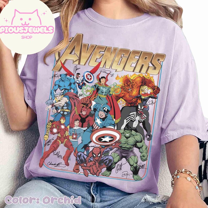 Puede incluir: Una camiseta de color orqu&iacute;dea con un gran estampado gr&aacute;fico de varios personajes de c&oacute;mics de Marvel, incluidos Spider-Man, Hulk e Iron Man, con la palabra AVENGERS en letras doradas grandes en la parte superior y varias firmas.