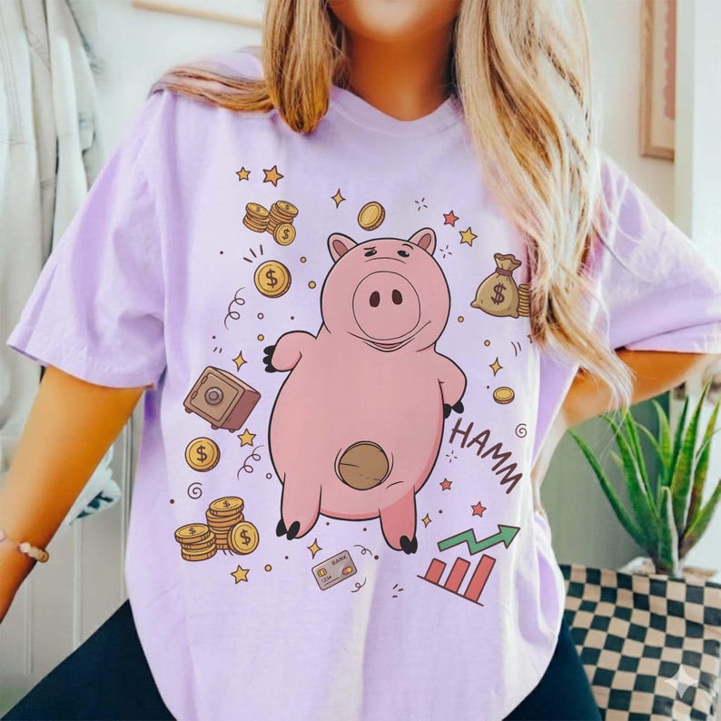 May include: A light purple t-shirt featuring a cartoon pig with the word "HAMM" in black. The design includes gold coins, stars, a safe, a money bag, a credit card, and a rising graph.