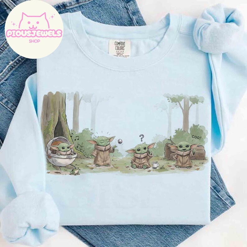 May include: Light blue crew neck sweatshirt featuring a cartoon scene with a green alien character in a forest setting. The design includes the text "Coo!" and a question mark. The sweatshirt has the "Comfort Colors" brand tag.