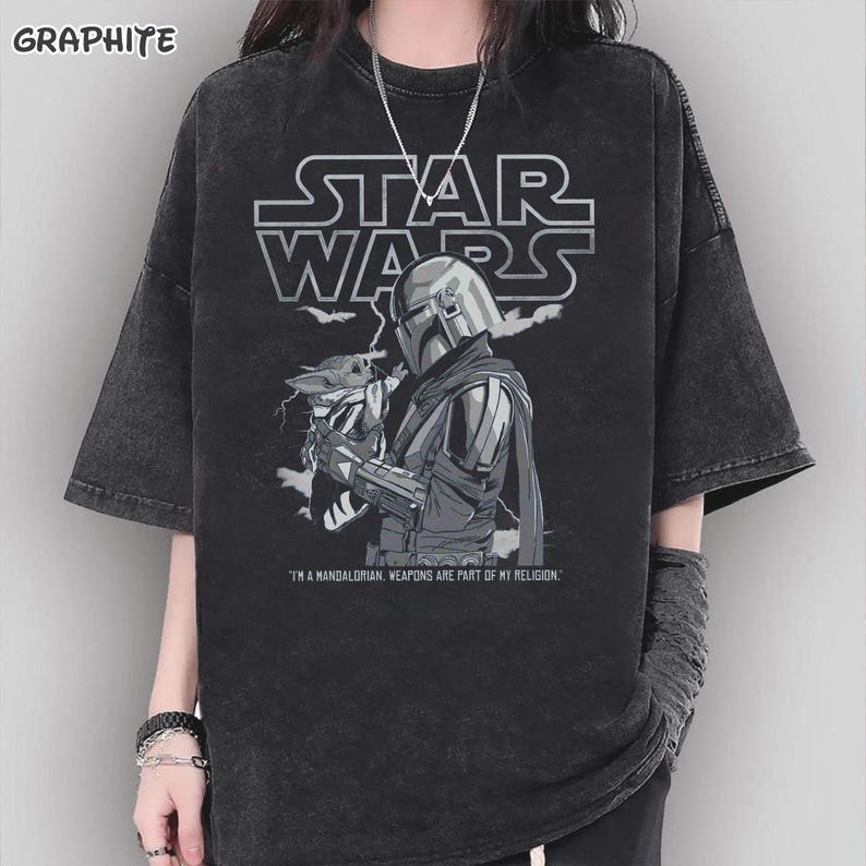 Op de afbeelding: Graphietkleurig Star Wars T-shirt met een afbeelding van The Mandalorian met Grogu. Het Star Wars-logo staat boven de afbeelding. De tekst "I'M A MANDALORIAN. WEAPONS ARE PART OF MY RELIGION." staat eronder.