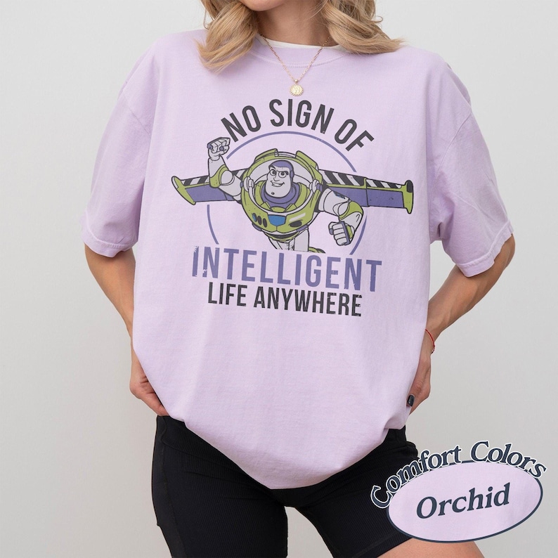 Pu&ograve; includere: T-shirt lilla chiaro con grafica di Buzz Lightyear e la scritta "NO SIGN OF INTELLIGENT LIFE ANYWHERE". La maglietta &egrave; un Comfort Colors Orchid. Il design presenta Buzz Lightyear in posa di volo.