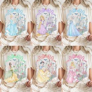 Op de afbeelding: Cr&egrave;mekleurige T-shirts met aquarel illustraties van Disney-prinsessen. Elk shirt toont een andere prinses in een jurk, met een kasteelachtergrond en bloemaccenten. Linten en strikken omlijsten de ontwerpen.