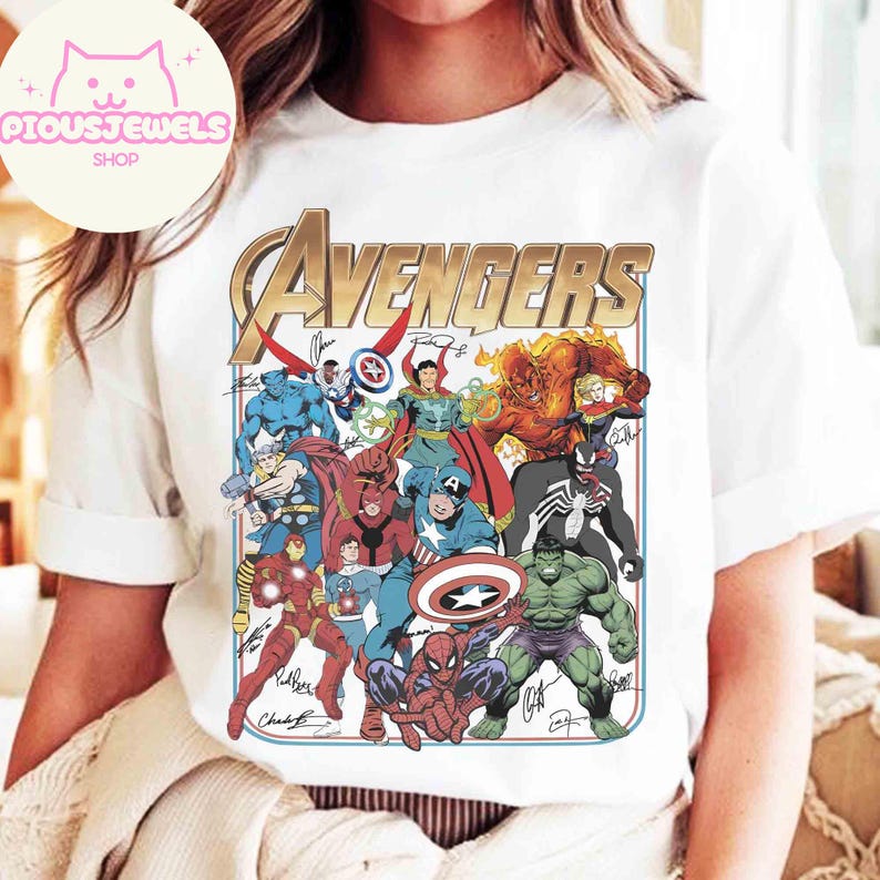 Puede incluir: Una camiseta blanca de manga corta con un gran estampado gr&aacute;fico de varios personajes de c&oacute;mics de Marvel, incluidos Spider-Man, Hulk, Capit&aacute;n Am&eacute;rica e Iron Man. La palabra AVENGERS est&aacute; impresa en letras doradas grandes sobre ellos.