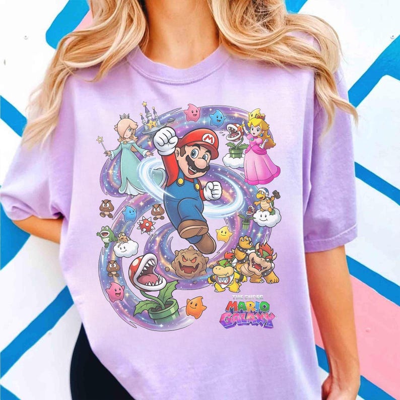 K&ouml;nnte beinhalten: Helllila T-Shirt mit einem farbenfrohen Aufdruck von Mario beim Springen, umgeben von Charakteren aus Super Mario Galaxy. Prinzessin Peach, Rosalina und verschiedene Feinde sind ebenfalls abgebildet. Der Text "THE CHEEP MARIO GALAXY" ist unten zu sehen.