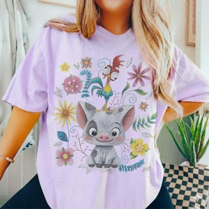 Puede incluir: Camiseta lila claro con un dise&ntilde;o de cerdo y gallo de dibujos animados, rodeados de flores. El cerdo es gris con orejas rosas y una cara sonriente. El gallo est&aacute; posado sobre la cabeza del cerdo. Flores coloridas.