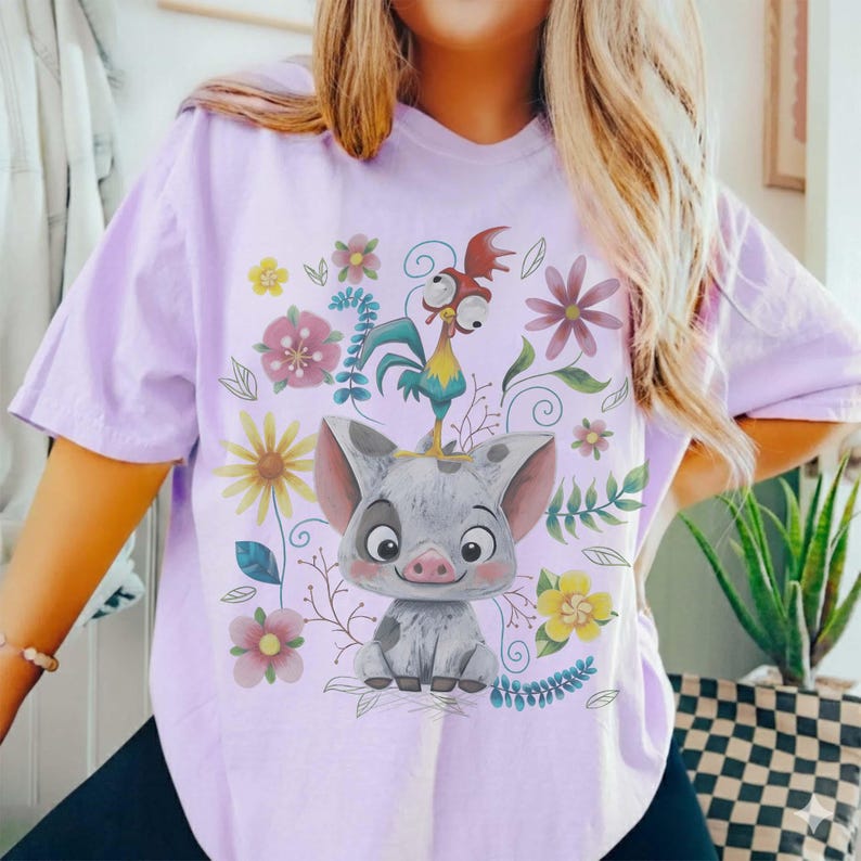 Puede incluir: Camiseta lila claro con un dise&ntilde;o de cerdo y gallo de dibujos animados, rodeados de flores. El cerdo es gris con orejas rosas y una cara sonriente. El gallo est&aacute; posado sobre la cabeza del cerdo. Flores coloridas.