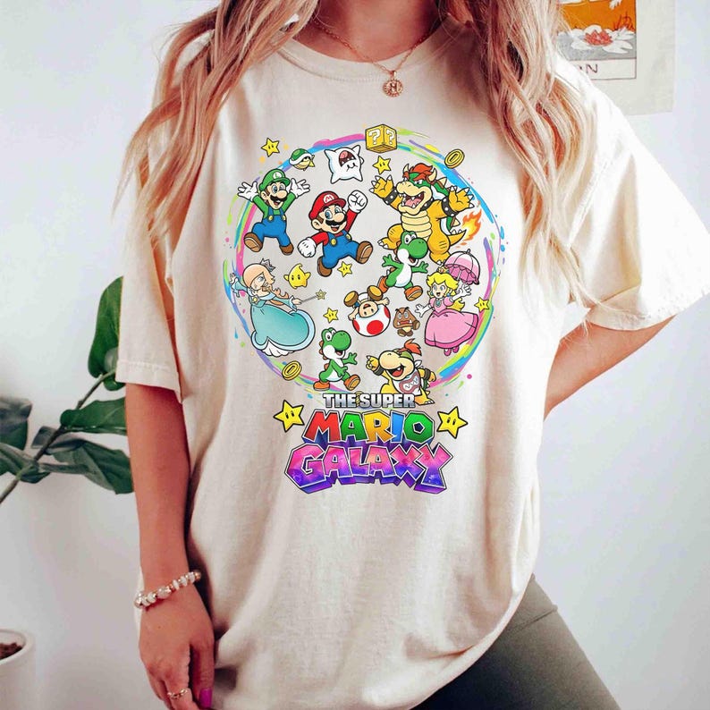 K&ouml;nnte beinhalten: Cremefarbenes T-Shirt mit einem farbenfrohen Grafikdesign von Charakteren aus dem Videospiel Super Mario Galaxy. Das Design zeigt Mario, Luigi, Prinzessin Peach und Bowser, mit dem Text "THE SUPER MARIO GALAXY".