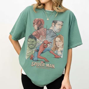 May include: Green t-shirt with a graphic of Spider-Man and other Marvel characters. The design includes Spider-Man in a crouching pose, surrounded by images of Daredevil, Hulk, and others. The text "SPIDER-MAN BRAND NEW DAY" is printed at the bottom.