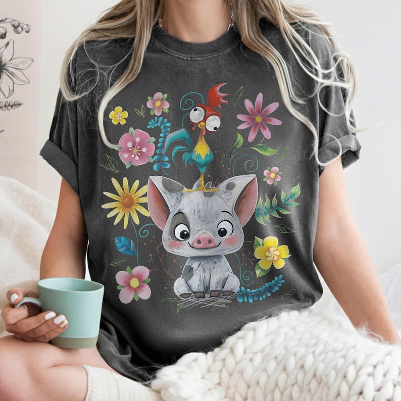Puede incluir: Camiseta gris oscuro con un cerdo de dibujos animados y un gallo, rodeados de flores coloridas. El dise&ntilde;o incluye elementos florales amarillos, rosas y azules. La camiseta tiene mangas cortas y un corte holgado.