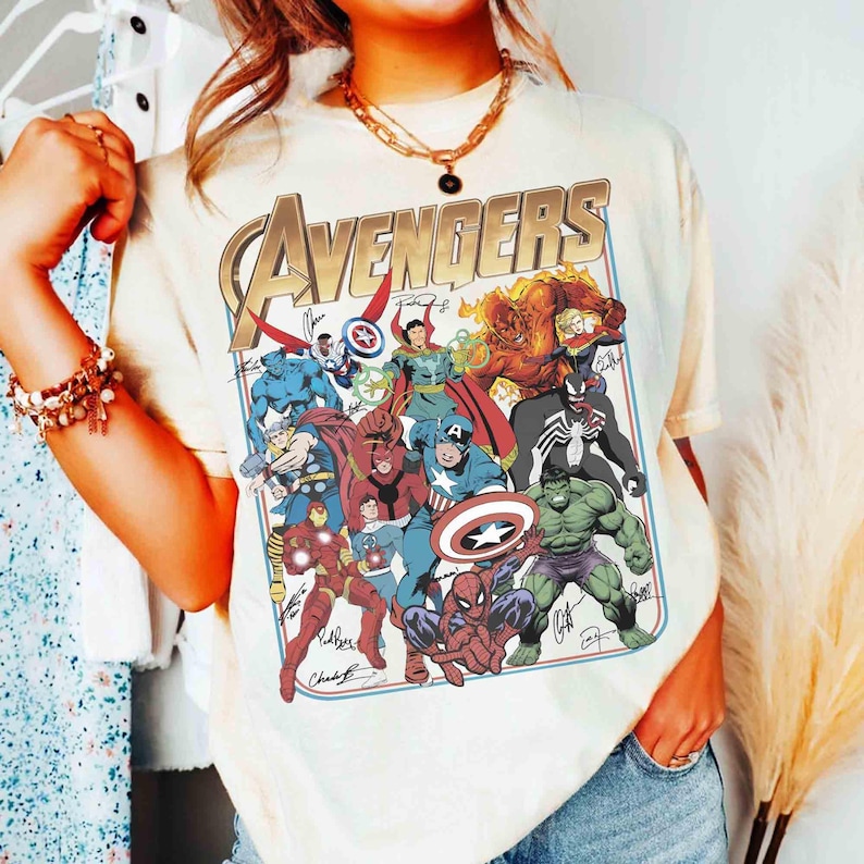 Puede incluir: Una camiseta de manga corta de color crema con un gran estampado gr&aacute;fico de varios personajes de c&oacute;mics de Marvel, incluidos Spider-Man, Hulk e Iron Man. La palabra AVENGERS est&aacute; impresa en letras doradas grandes en el dise&ntilde;o.