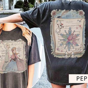Pode incluir: T-shirt cinza escuro com um gr&aacute;fico do filme Frozen. A frente mostra Elsa e Anna, enquanto a parte de tr&aacute;s exibe partituras com Olaf e um floco de neve. O texto "Let It Go" &eacute; vis&iacute;vel. Uma camiseta casual e confort&aacute;vel.