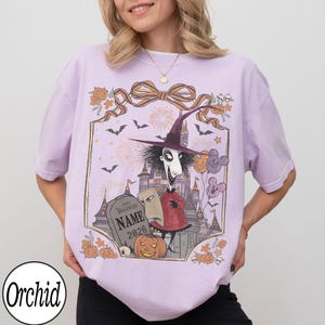 K&ouml;nnte beinhalten: Helllila T-Shirt mit einem Halloween-Motiv. Das Design zeigt eine Figur mit Hexenhut, ein Schloss, einen Grabstein und einen K&uuml;rbis. Der Text "Happy Halloween" und "Name 2026" sind auf dem Grabstein zu sehen. Das Shirt hat auch den Markennamen "Orchid".