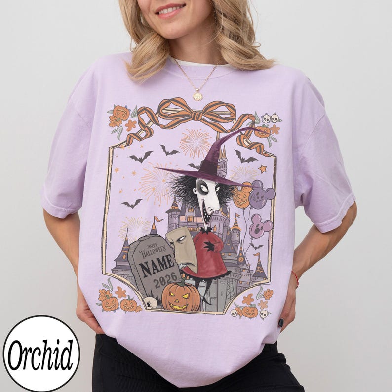 K&ouml;nnte beinhalten: Helllila T-Shirt mit einem Halloween-Motiv. Das Design zeigt eine Figur mit Hexenhut, ein Schloss, einen Grabstein und einen K&uuml;rbis. Der Text "Happy Halloween" und "Name 2026" sind auf dem Grabstein zu sehen. Das Shirt hat auch den Markennamen "Orchid".