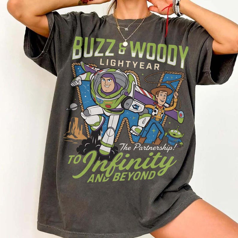 Może przedstawiać: Ciemnoszara koszulka z nadrukiem przedstawiającym Buzza Astrala i Chudego z Toy Story. Grafika zawiera tekst "Buzz & Woody Lightyear" i "To Infinity and Beyond". Projekt przedstawia postacie na tle pustynnego krajobrazu.