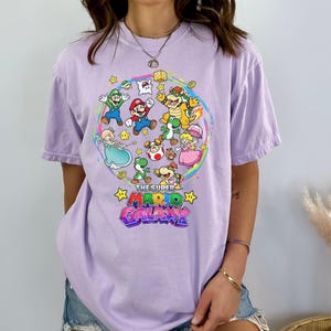 K&ouml;nnte beinhalten: Helllila T-Shirt mit farbenfroher Grafik von Charakteren aus Super Mario Galaxy. Das Design zeigt Mario, Luigi, Prinzessin Peach, Yoshi und mehr, vor einem Regenbogenhintergrund. Der Titel des Spiels ist im Sternstil dargestellt.