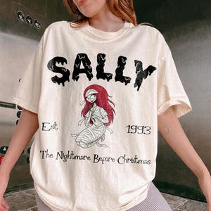 K&ouml;nnte beinhalten: Cremefarbenes T-Shirt mit dem Wort "SALLY" in schwarzer Tropfschrift. Darunter ist eine Illustration von Sally aus "The Nightmare Before Christmas" abgebildet. Der Text "Est 1993 The Nightmare Before Christmas" ist ebenfalls aufgedruckt.