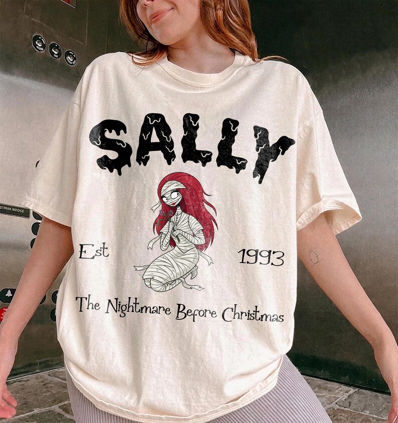 K&ouml;nnte beinhalten: Cremefarbenes T-Shirt mit dem Wort "SALLY" in schwarzer Tropfschrift. Darunter ist eine Illustration von Sally aus "The Nightmare Before Christmas" abgebildet. Der Text "Est 1993 The Nightmare Before Christmas" ist ebenfalls aufgedruckt.