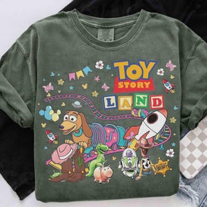 May include: Green crew neck sweatshirt with a colourful graphic of Toy Story characters and the words "Toy Story Land." The design includes a rocket, a train, and various characters like Buzz Lightyear and Woody. The sweatshirt has a vintage, washed look.