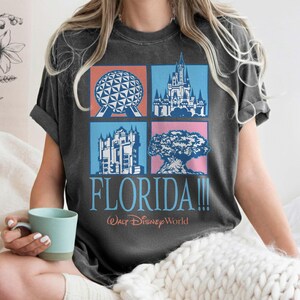 May include: Dark gray t-shirt featuring a colorful graphic of Florida landmarks, including a sphere, castle, and tree. The word "FLORIDA!!!" is printed below the images, with "Walt Disney World" underneath.